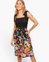 A woman modeling a sleeveless midi dress with a black smocked bodice and a black skirt featuring a vibrant multicolor floral print, set against a plain white background.