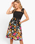 A woman modeling a sleeveless midi dress with a black smocked bodice and a vibrant multicolor floral skirt, set against a plain light gray background.