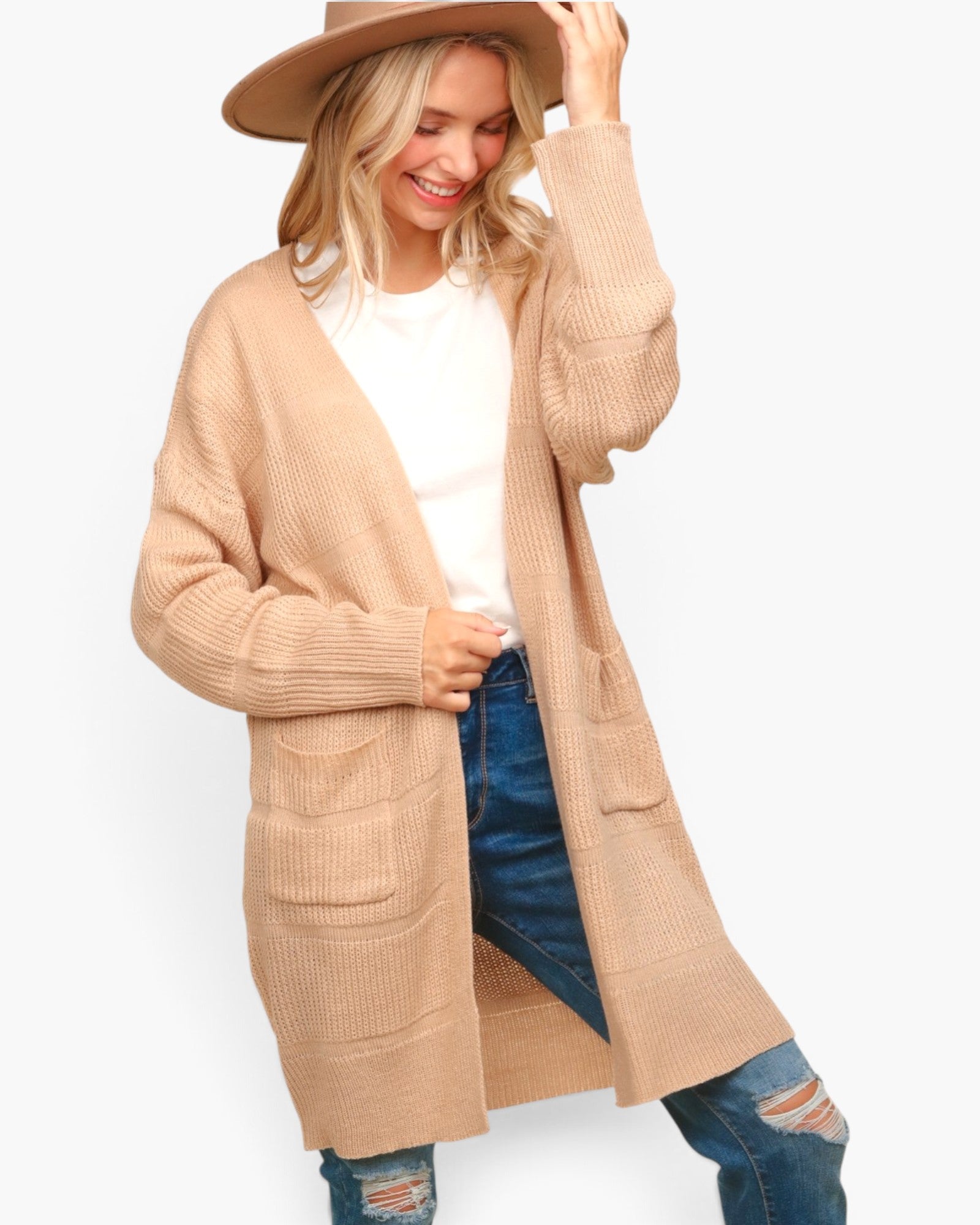 A woman wearing a long beige ribbed knit cardigan over a white top and blue jeans, with a matching beige wide-brim hat, photographed against a light gray studio background.