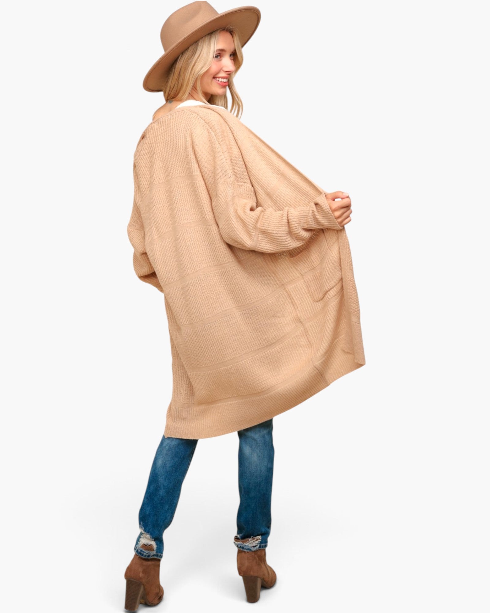 A woman wearing a light beige oversized knit cardigan, blue jeans, brown ankle boots, and a matching wide-brim hat, photographed against a plain light gray studio background.