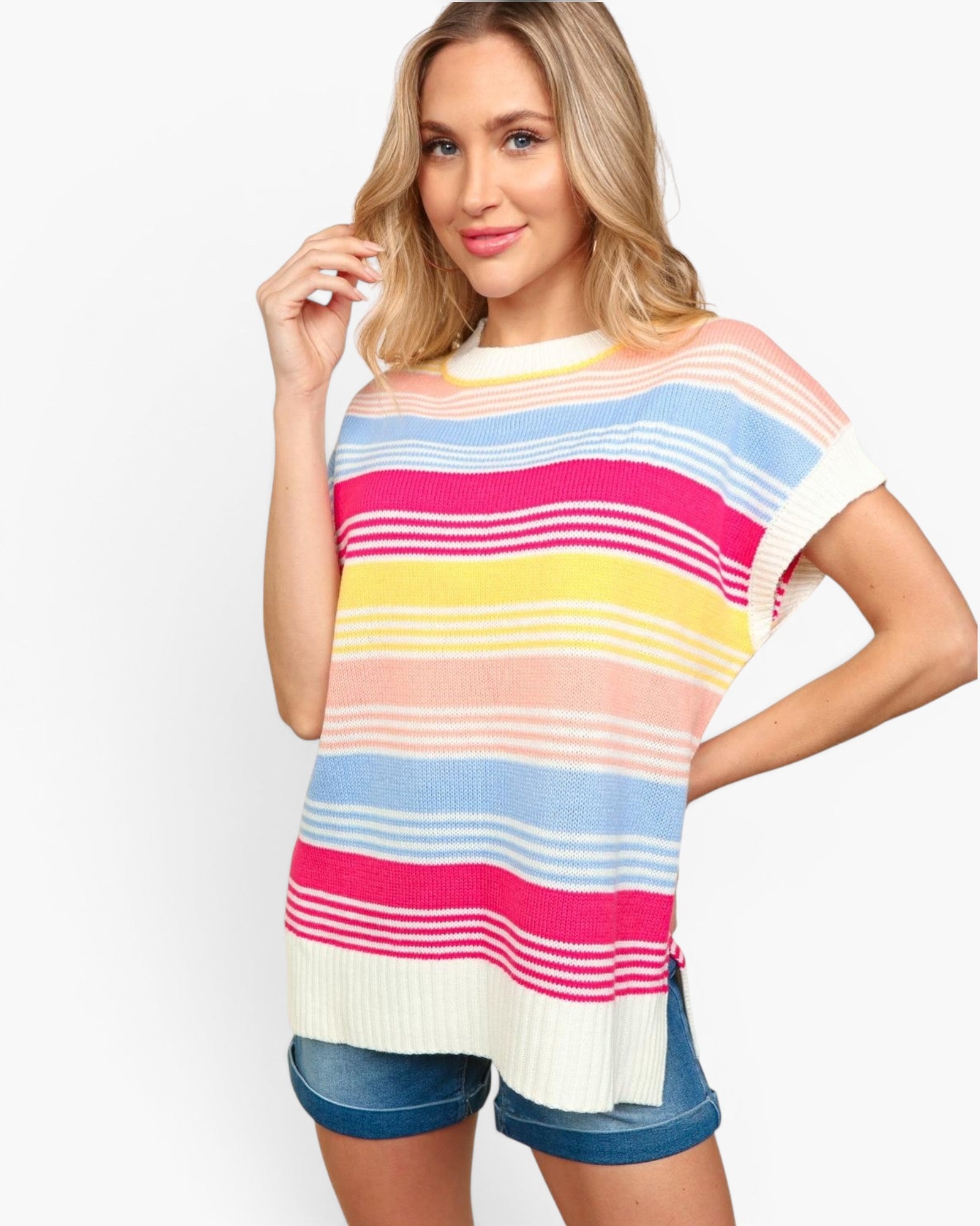 A woman wearing a colorful striped knit tunic sweater with short sleeves and denim shorts, posed against a clean white studio background.