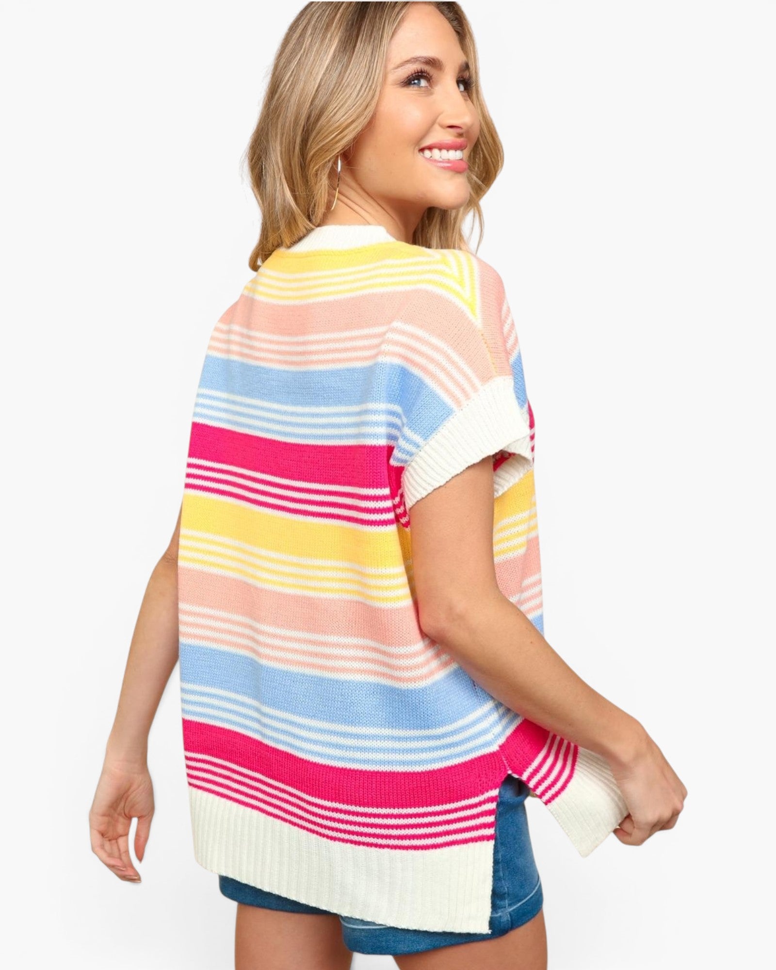 A woman wearing a colorful striped knit poncho-style top with horizontal bands of pink, yellow, light blue, peach, and white, shown from a three-quarter back view against a plain light gray studio background.