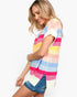 A woman wearing a colorful striped knit top with pink, blue, yellow, and white horizontal stripes, paired with denim shorts, against a plain white background.