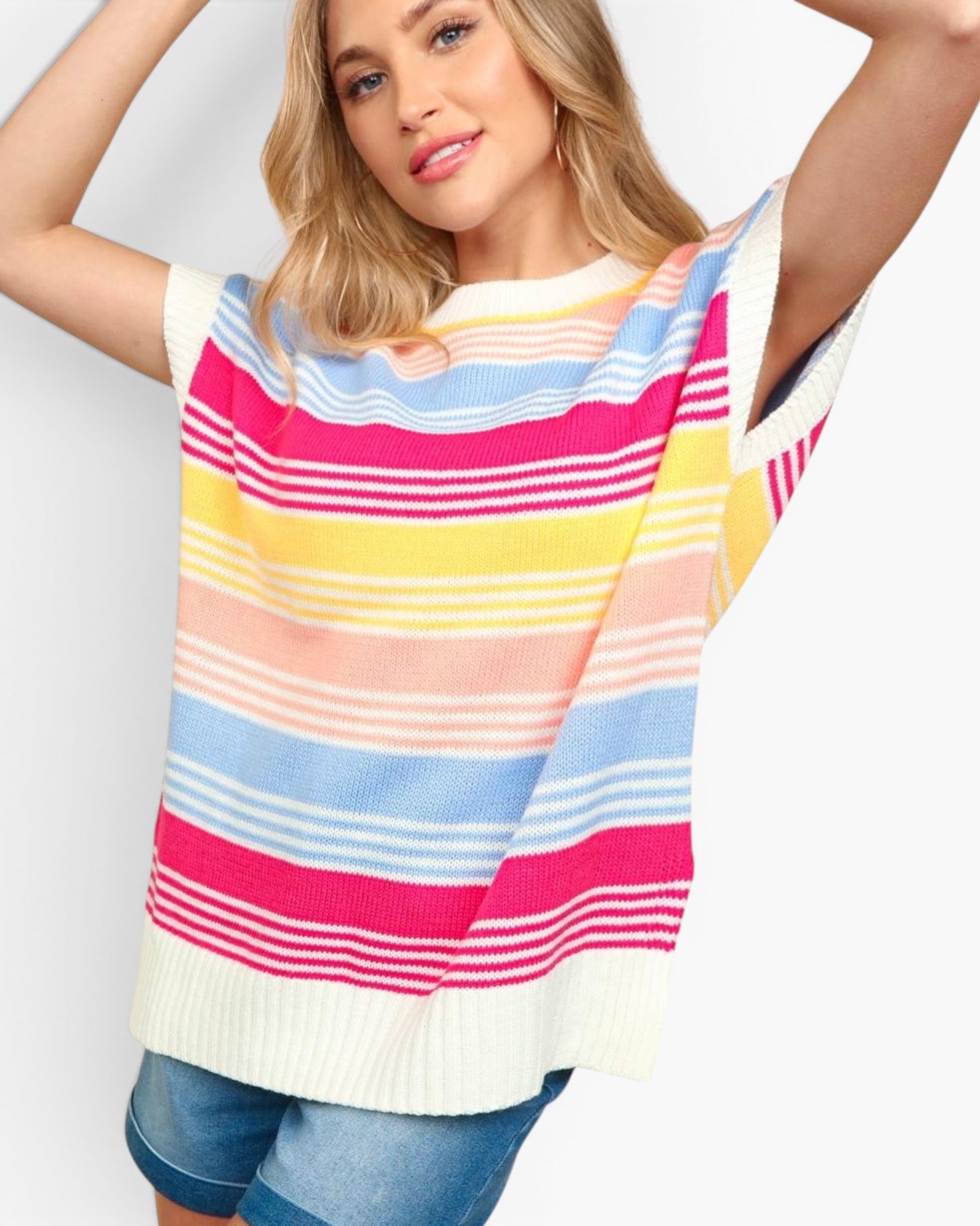 A woman wearing a colorful striped knit top with horizontal bands of pink, yellow, light blue, peach, and white, paired with blue denim shorts, against a plain light gray background.
