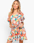 A woman modeling a short-sleeve wrap-style dress with a tie waist and a vibrant tropical floral print in orange, white, teal, and yellow against a plain light gray studio background.