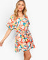 A woman modeling a short tropical floral print dress with short sleeves, a V-neck, and a tie waist, set against a plain white background.