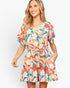 A woman modeling a short-sleeve floral romper with a tie waist, featuring a vibrant print of orange, teal, white, and yellow flowers on a light background, set against a plain light gray studio backdrop.