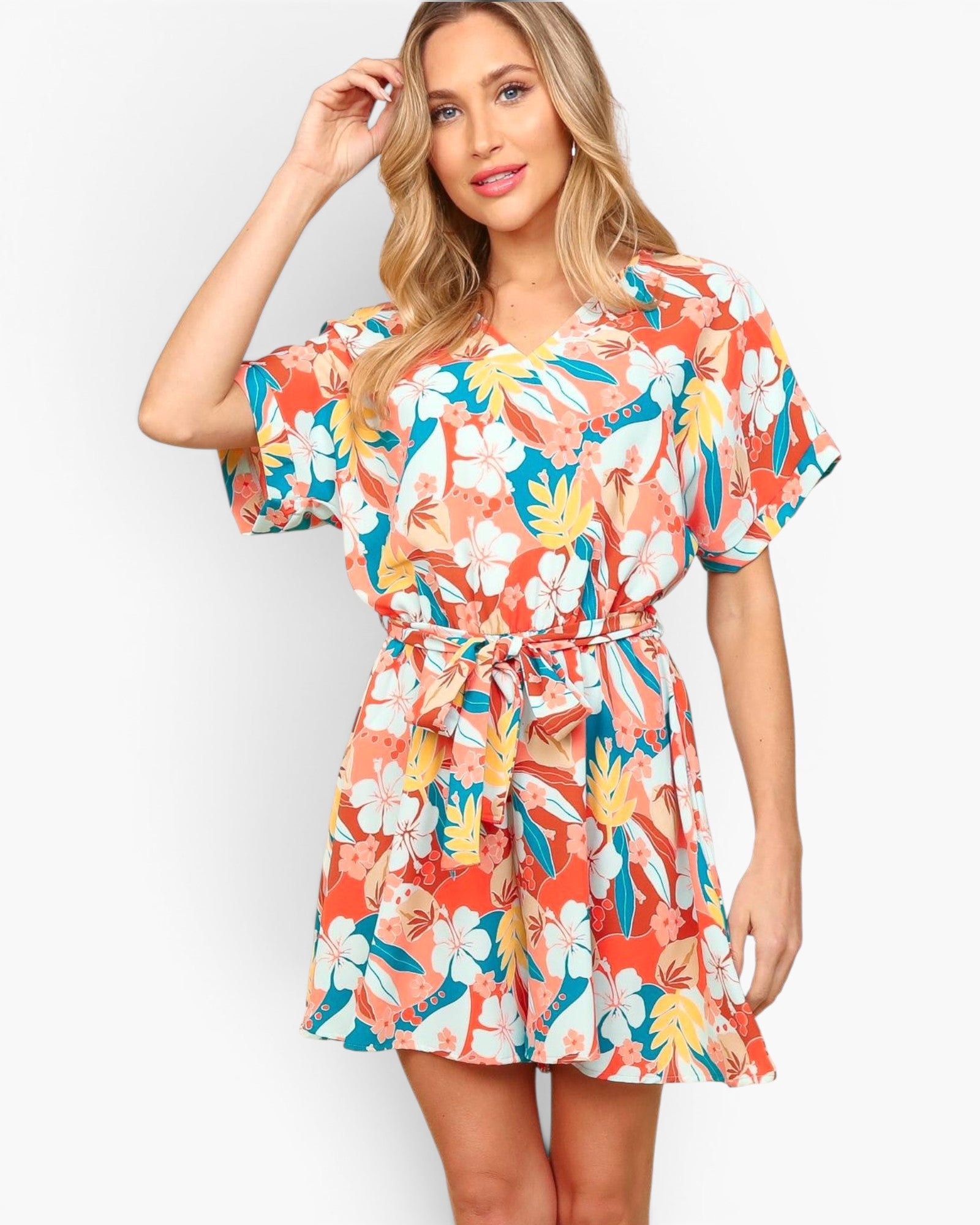 A woman modeling a short-sleeve V-neck mini dress with a colorful tropical floral print in orange, teal, yellow, and white, set against a plain light gray background.