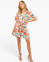 A woman modeling a short-sleeve floral romper with a tie waist, featuring a vibrant orange, pink, blue, and white tropical pattern, set against a plain white studio background.