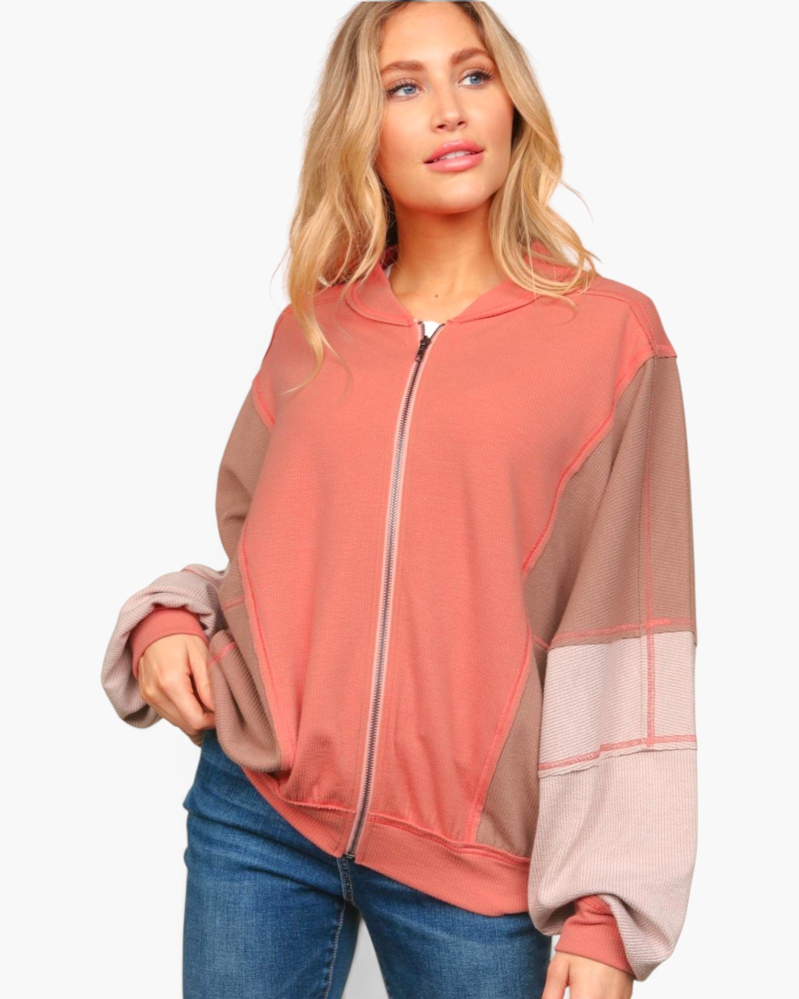 A woman wearing a color-blocked zip-up sweatshirt with a dusty rose front panel, taupe shoulder panels, and light pink sleeve panels, paired with blue jeans against a plain white background.