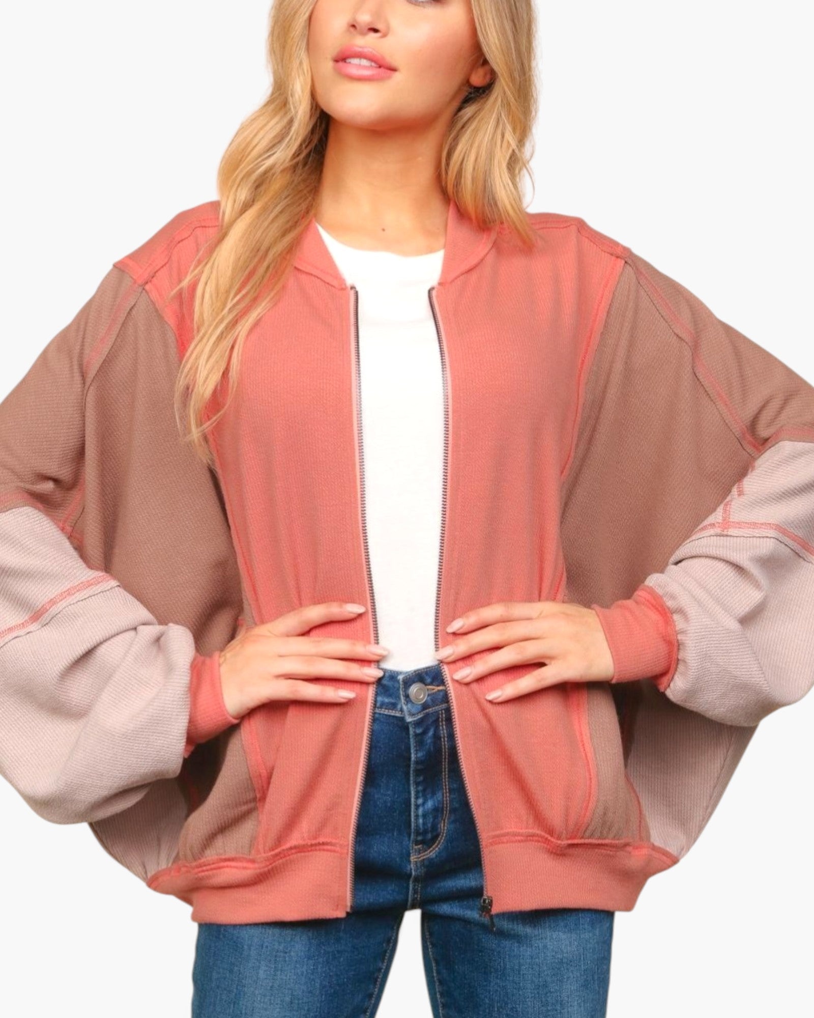 A woman wearing a color-blocked bomber jacket with a dusty rose front panel and sleeves, and taupe-brown shoulder panels, styled over a white t-shirt and blue jeans.