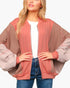 A woman wearing a color-blocked bomber jacket with a dusty rose front panel and sleeves, and taupe-brown shoulder panels, styled over a white t-shirt and blue jeans.