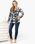 A woman modeling a black and white geometric patterned long-sleeve top with a drawstring neckline, paired with dark blue jeans, standing next to a white dresser against a white studio background.