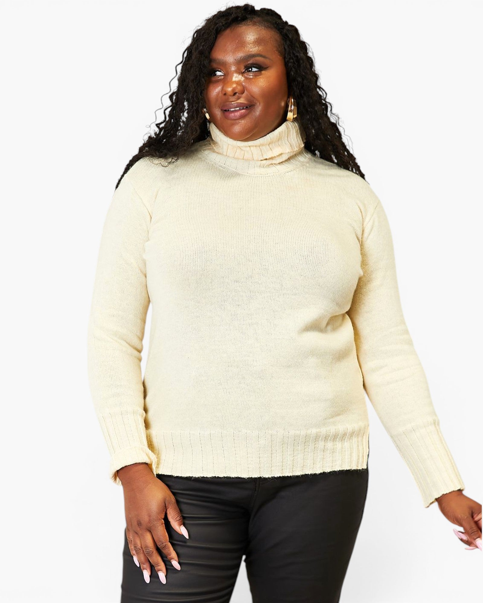 A woman wearing a cream-colored ribbed turtleneck sweater and black pants, standing against a plain white studio background.