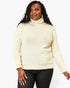 A woman wearing a cream-colored ribbed turtleneck sweater and black pants, standing against a plain white studio background.