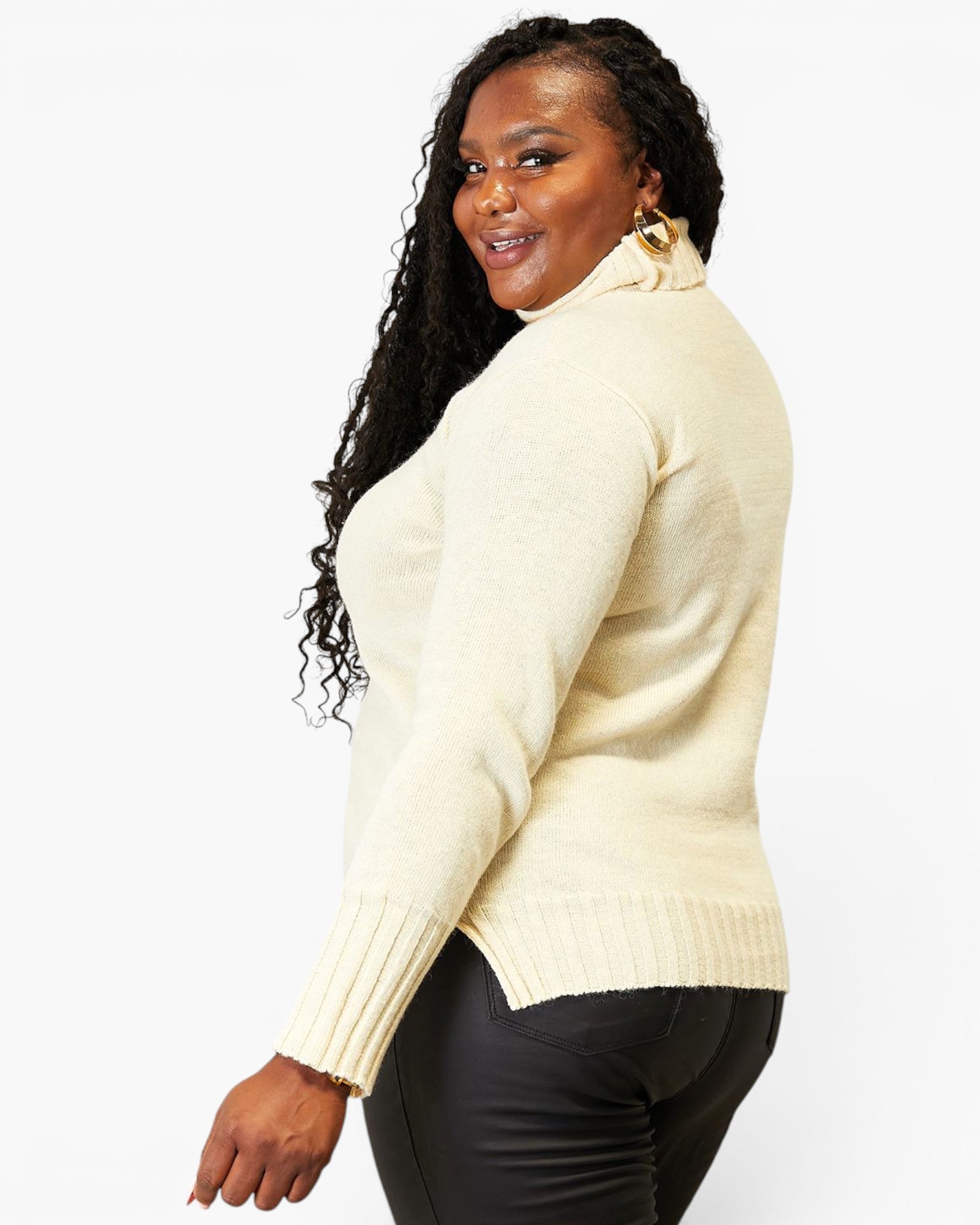 A woman wearing a cream-colored ribbed turtleneck sweater and black pants, shown from a side-back angle against a plain white studio background.