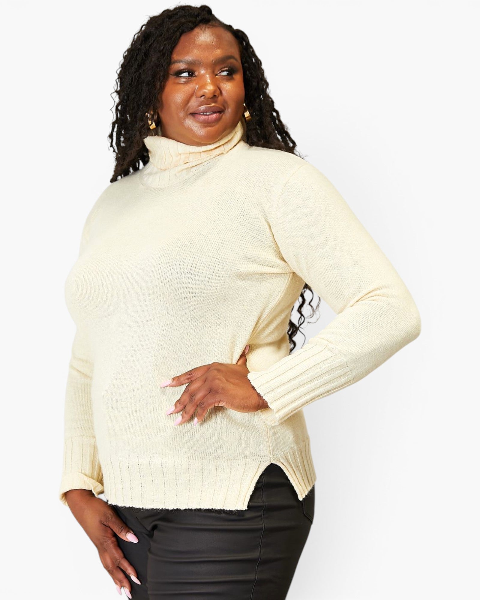 A woman modeling a cream-colored ribbed turtleneck sweater with black pants, photographed against a solid light gray background.