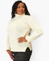 A woman modeling a cream-colored ribbed turtleneck sweater with black pants, photographed against a solid light gray background.
