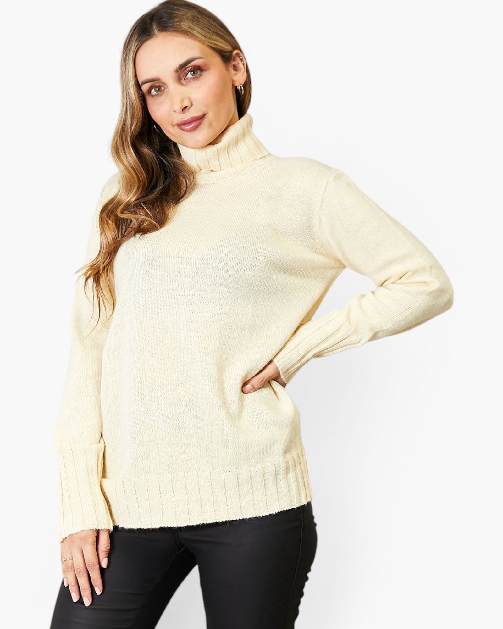 A woman wearing a cream-colored ribbed turtleneck sweater and black pants, posing against a light gray studio background.