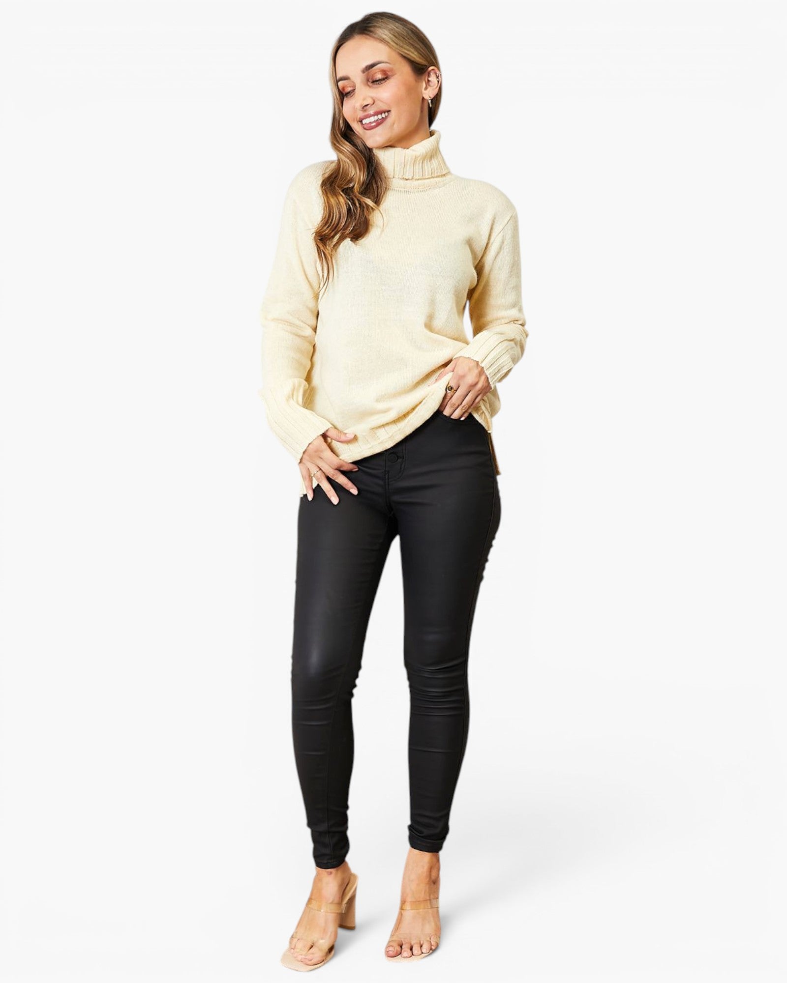 A woman modeling a cream-colored mock neck knit sweater and black skinny jeans against a plain white studio background.