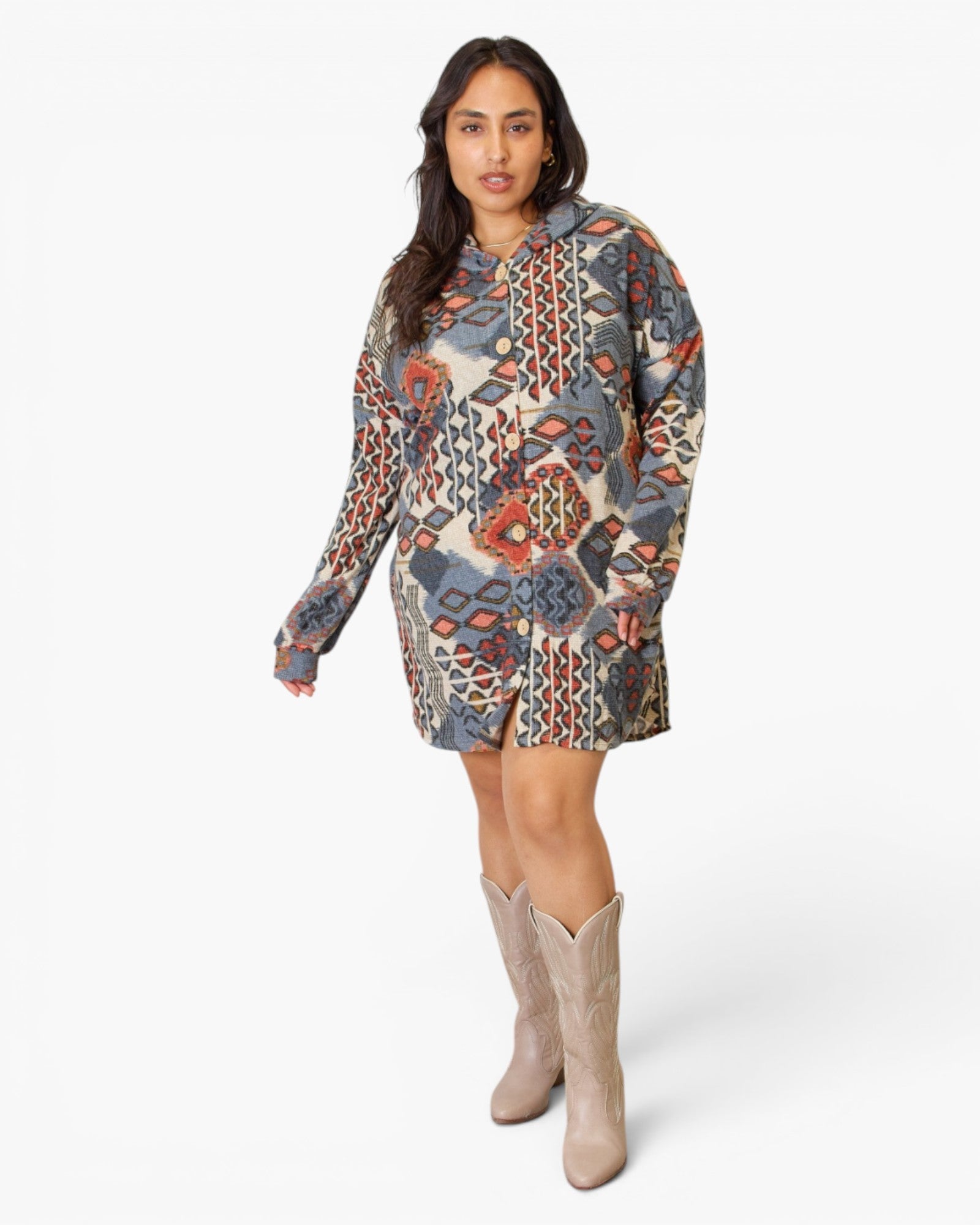 A woman modeling a long-sleeve hooded mini dress with a busy geometric and tribal-inspired print in muted blue, rust orange, beige, and gray tones, paired with beige cowboy boots against a light gray studio background.