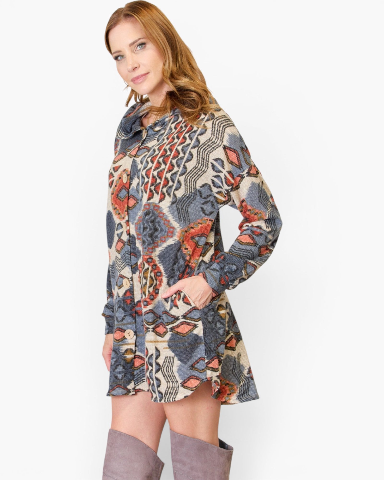 A woman modeling a long-sleeve hooded tunic dress with an abstract geometric pattern in beige, gray, blue, and rust orange, paired with gray knee-high boots against a plain white background.