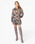 A woman modeling a long-sleeve hooded mini dress with a colorful geometric tribal pattern in blue, red, orange, and beige, paired with over-the-knee taupe boots, against a plain white studio background.