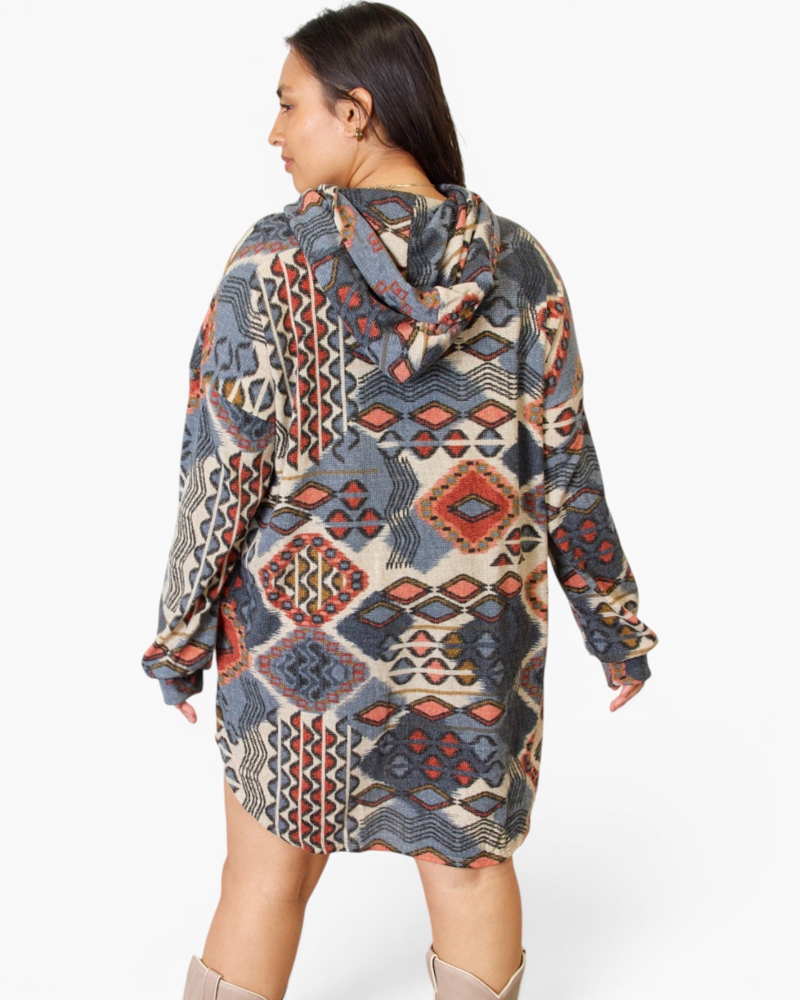 A woman shown from the back wearing a hooded long-sleeve tunic dress with a multicolor geometric tribal pattern in beige, blue, gray, and rust red, paired with beige boots against a light gray studio background.