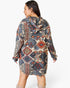 A woman shown from the back wearing a hooded long-sleeve tunic dress with a multicolor geometric tribal pattern in beige, blue, gray, and rust red, paired with beige boots against a light gray studio background.