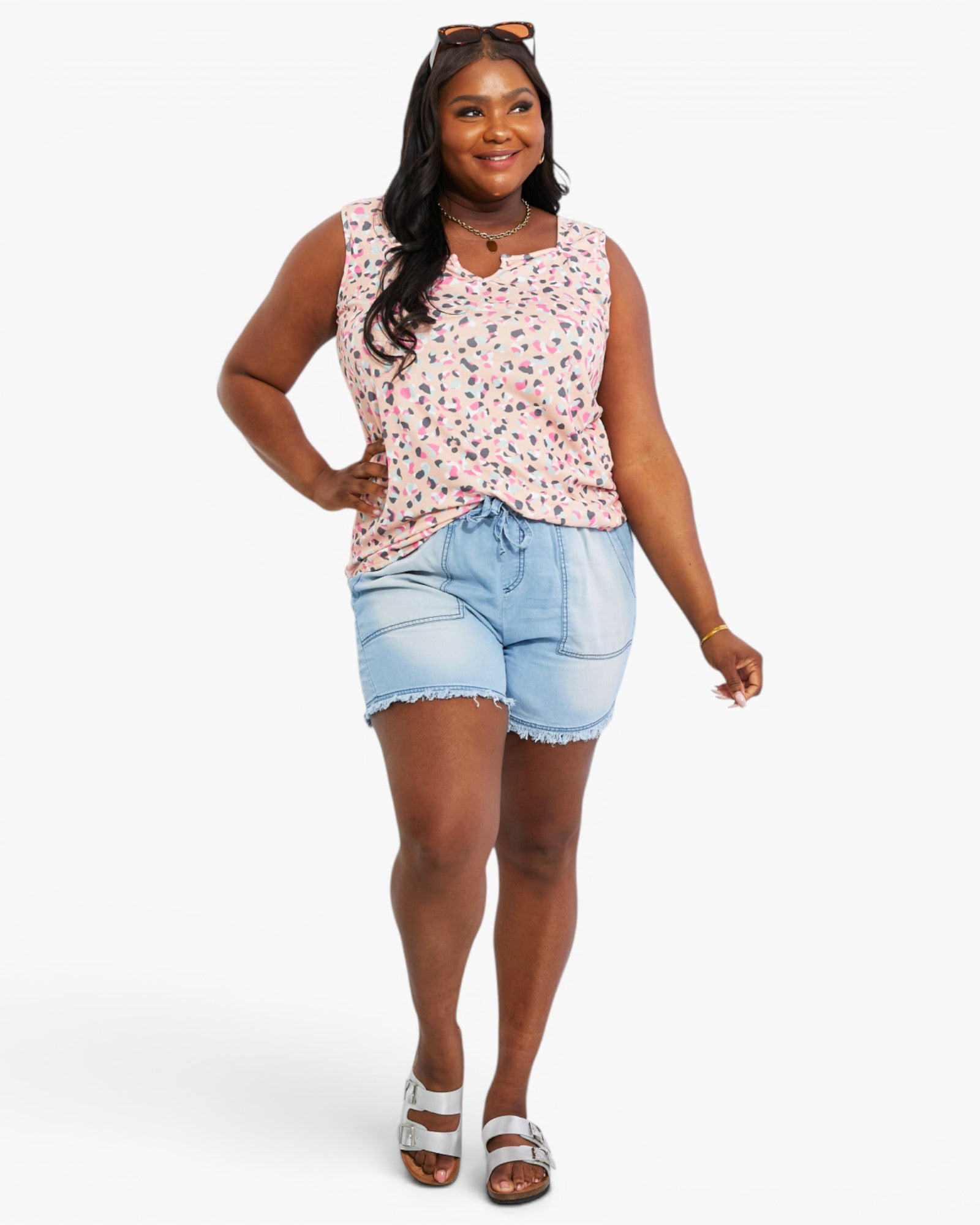 A woman modeling a sleeveless pink floral top and light blue denim shorts against a plain light gray studio background.