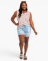 A woman modeling a sleeveless pink floral top and light blue denim shorts against a plain light gray studio background.