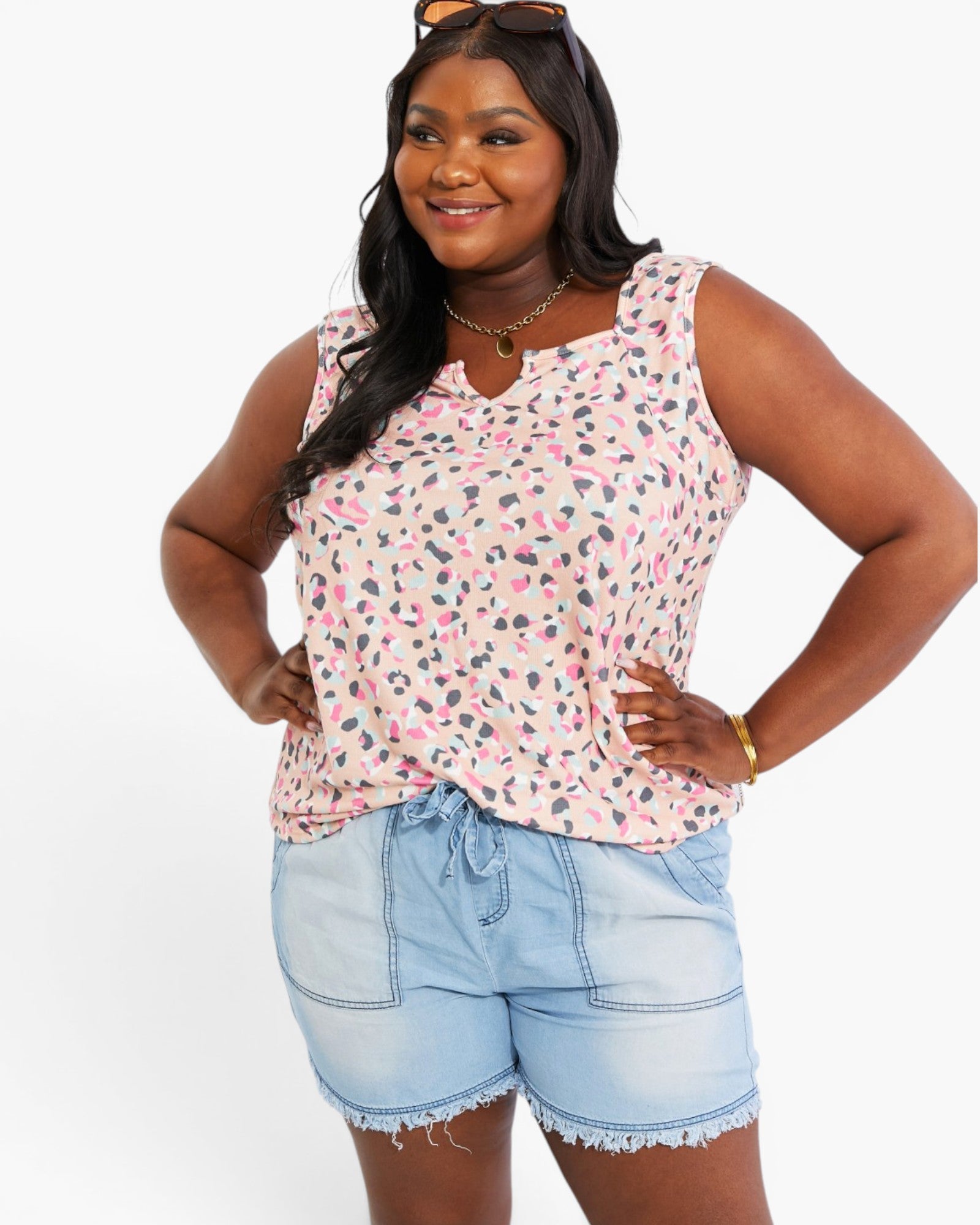 A woman modeling a sleeveless top with a pink, gray, and black leopard print on a white background, paired with light blue denim shorts, against a plain white studio background.