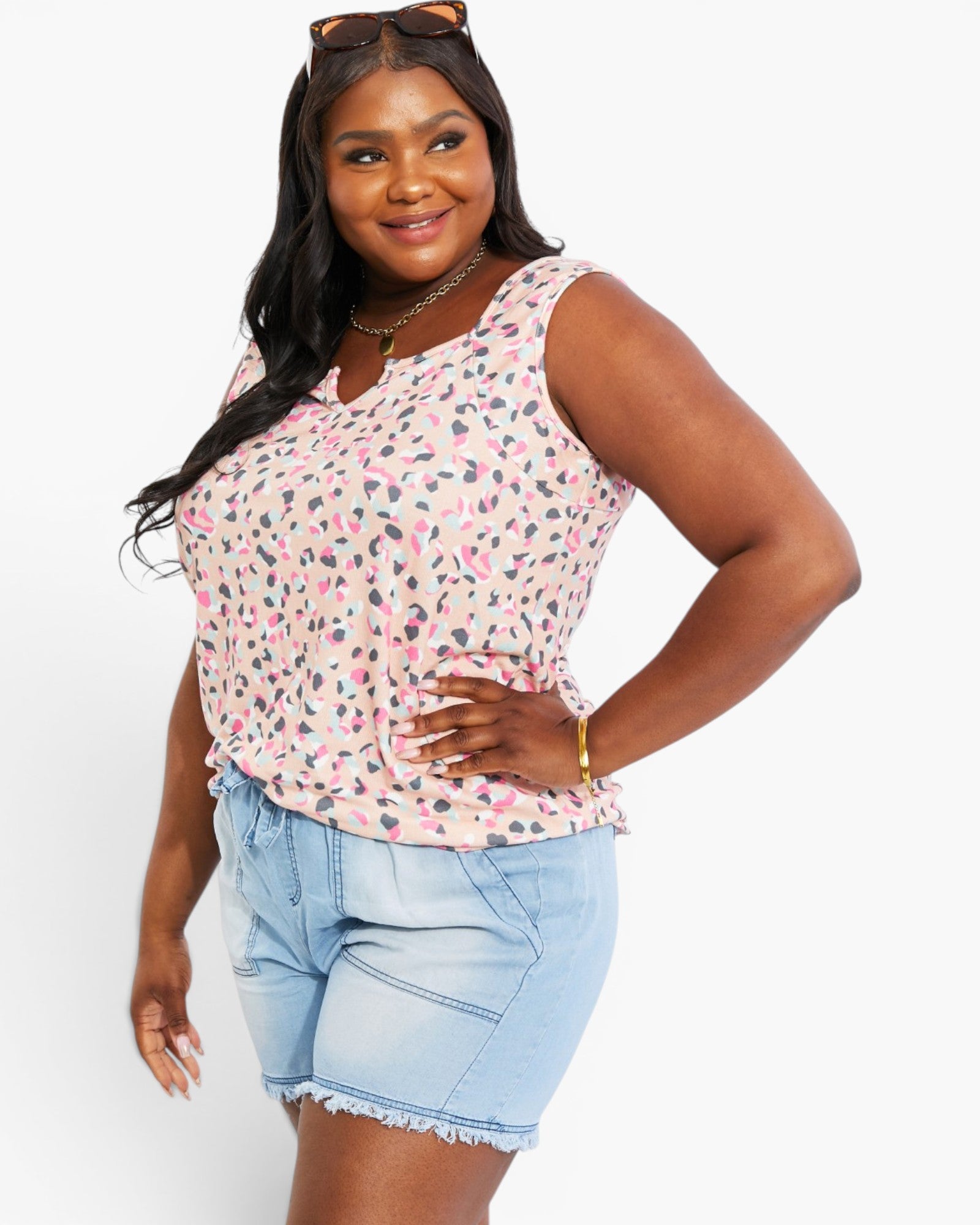 A woman modeling a sleeveless V-neck top with a pink, gray, and black leopard print pattern, paired with light blue denim shorts, against a plain white background.