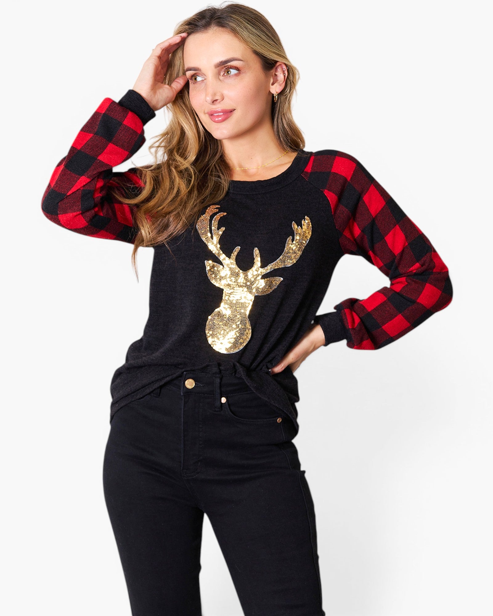 A woman modeling a long-sleeve top with a black and red buffalo plaid pattern on the sleeves and collar, paired with a black body featuring a large gold sequined deer head graphic on the chest, styled with black jeans against a plain white background.