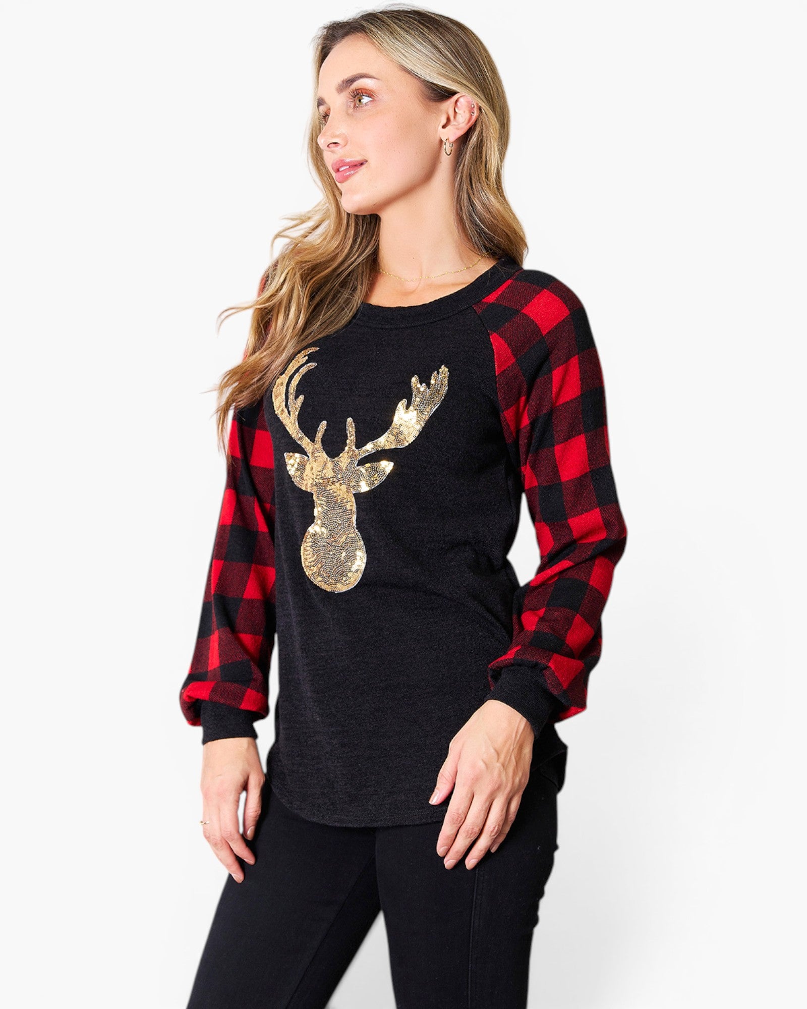 A woman wearing a long-sleeve holiday top with a black body, red and black buffalo plaid sleeves, and a large gold sequined deer graphic on the front, paired with black pants against a light gray studio background.