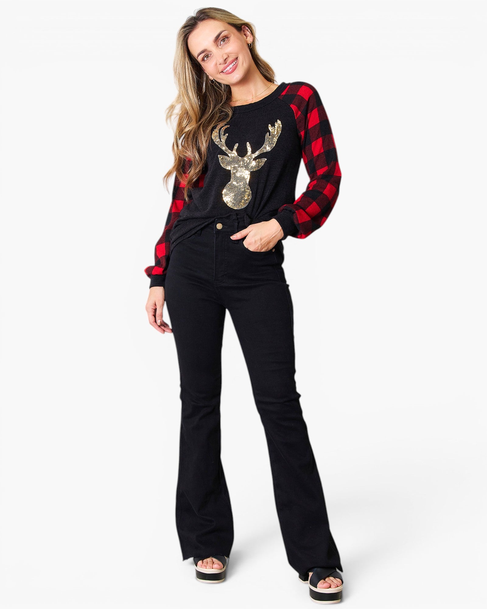 A woman modeling a black long-sleeve top with red buffalo plaid sleeves, a gold sequined reindeer graphic on the front, and black flared jeans against a plain white background.
