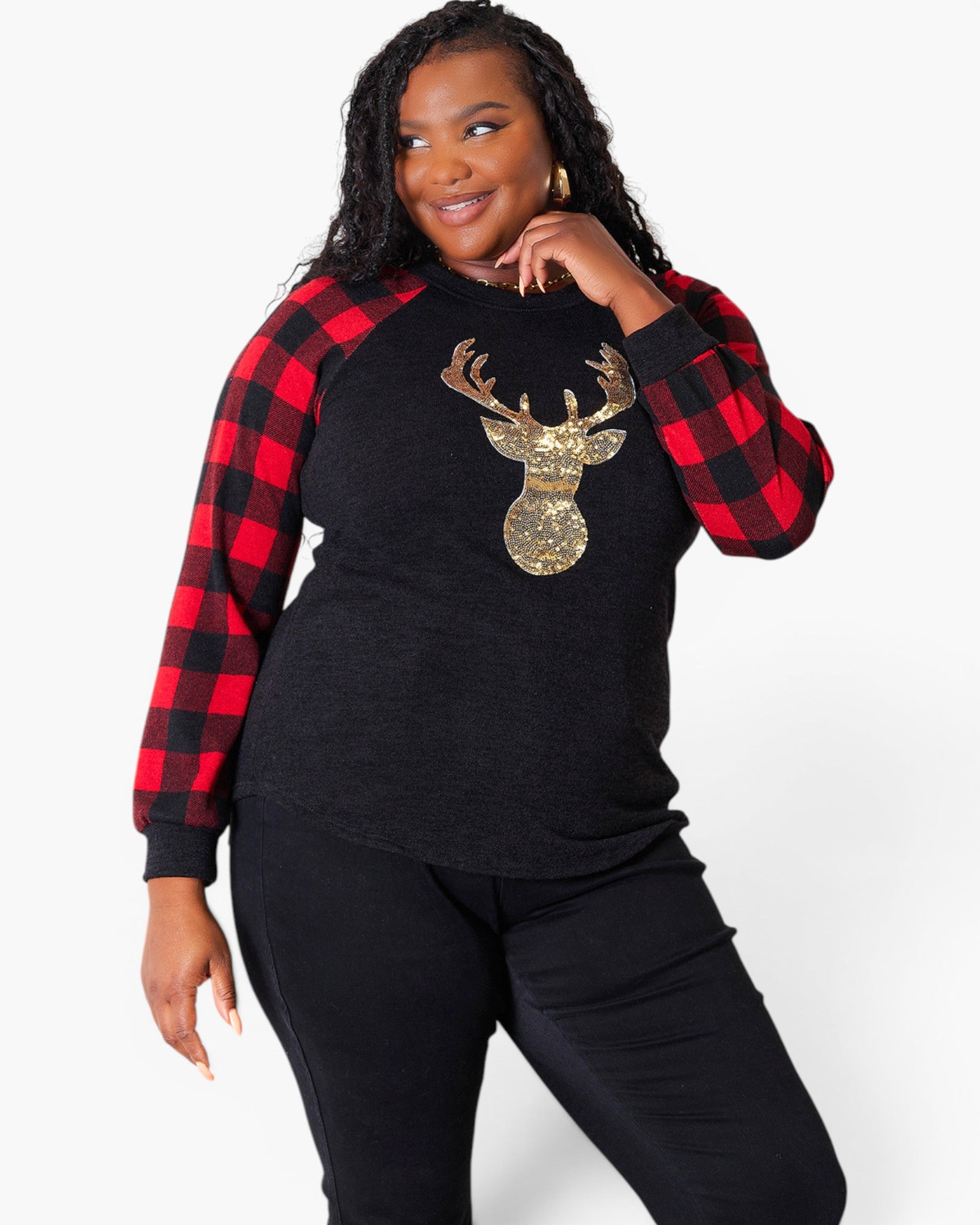 A woman modeling a black long-sleeve top with red and black buffalo plaid raglan sleeves and a large gold sequined deer head graphic on the front, paired with black pants against a light gray studio background.