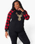A woman modeling a black long-sleeve top with red and black buffalo plaid raglan sleeves and a large gold sequined deer head graphic on the front, paired with black pants against a light gray studio background.