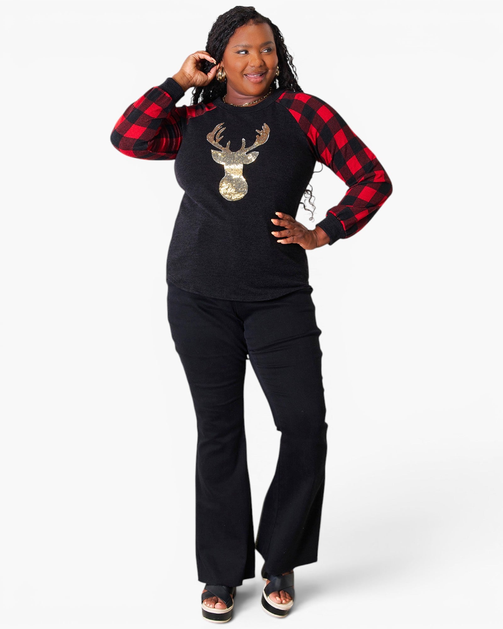 A woman modeling a black raglan long-sleeve top with red buffalo plaid sleeves, a gold sequined deer graphic on the chest, and black flared pants against a light gray background.