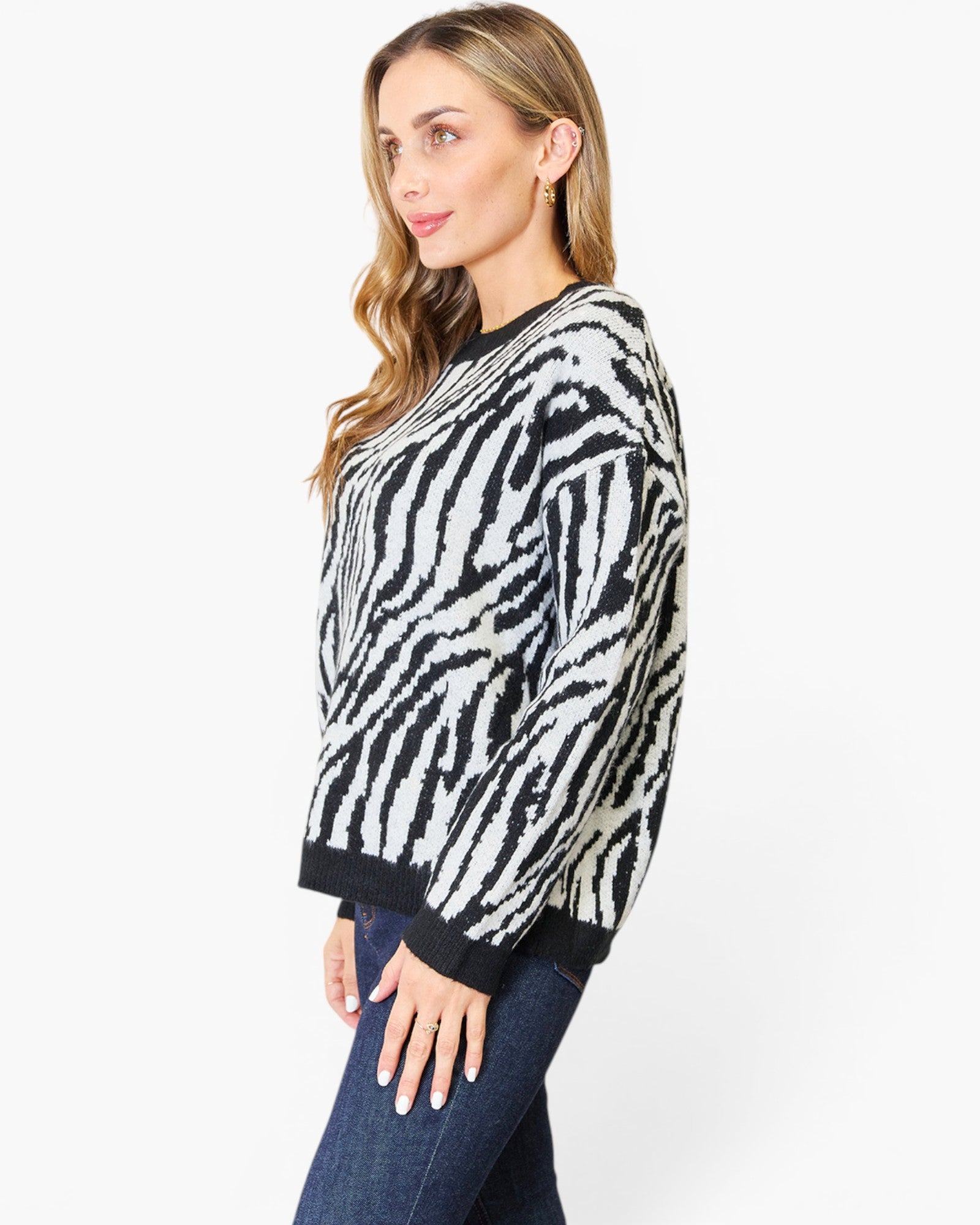 A woman wearing a black and white zebra-patterned knit sweater with dark ribbed trim, paired with dark blue jeans, photographed against a plain light gray studio background.
