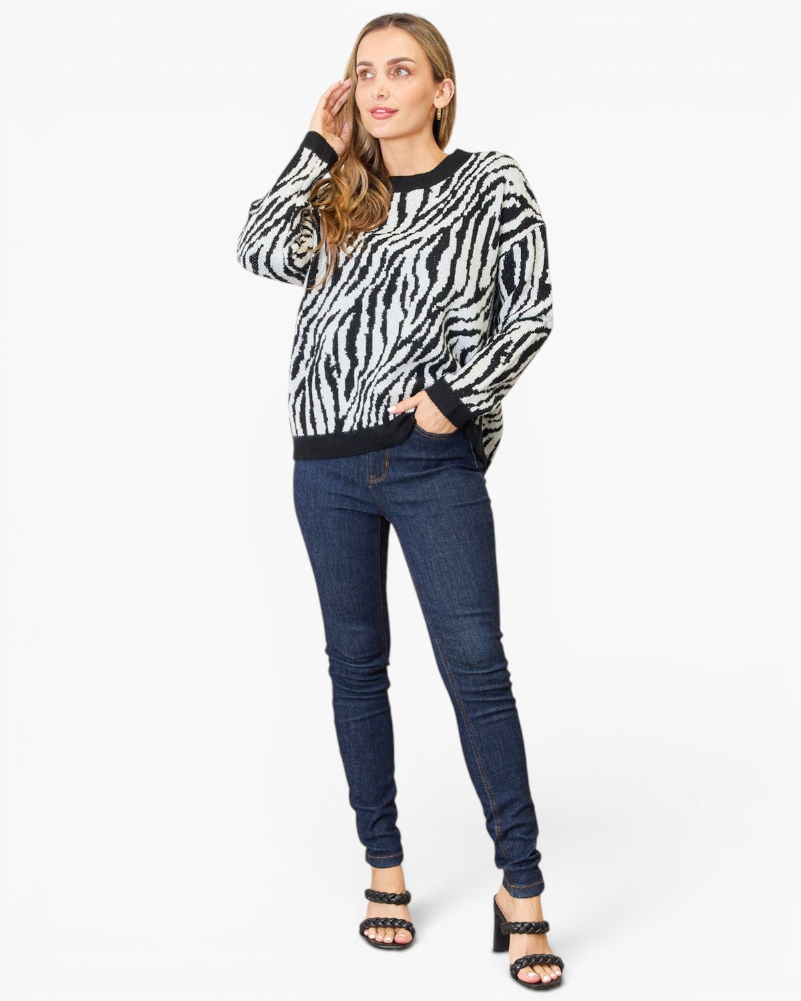 A woman modeling a black and white zebra-print long-sleeve sweater with dark blue skinny jeans and black heeled sandals, standing against a plain white studio background.