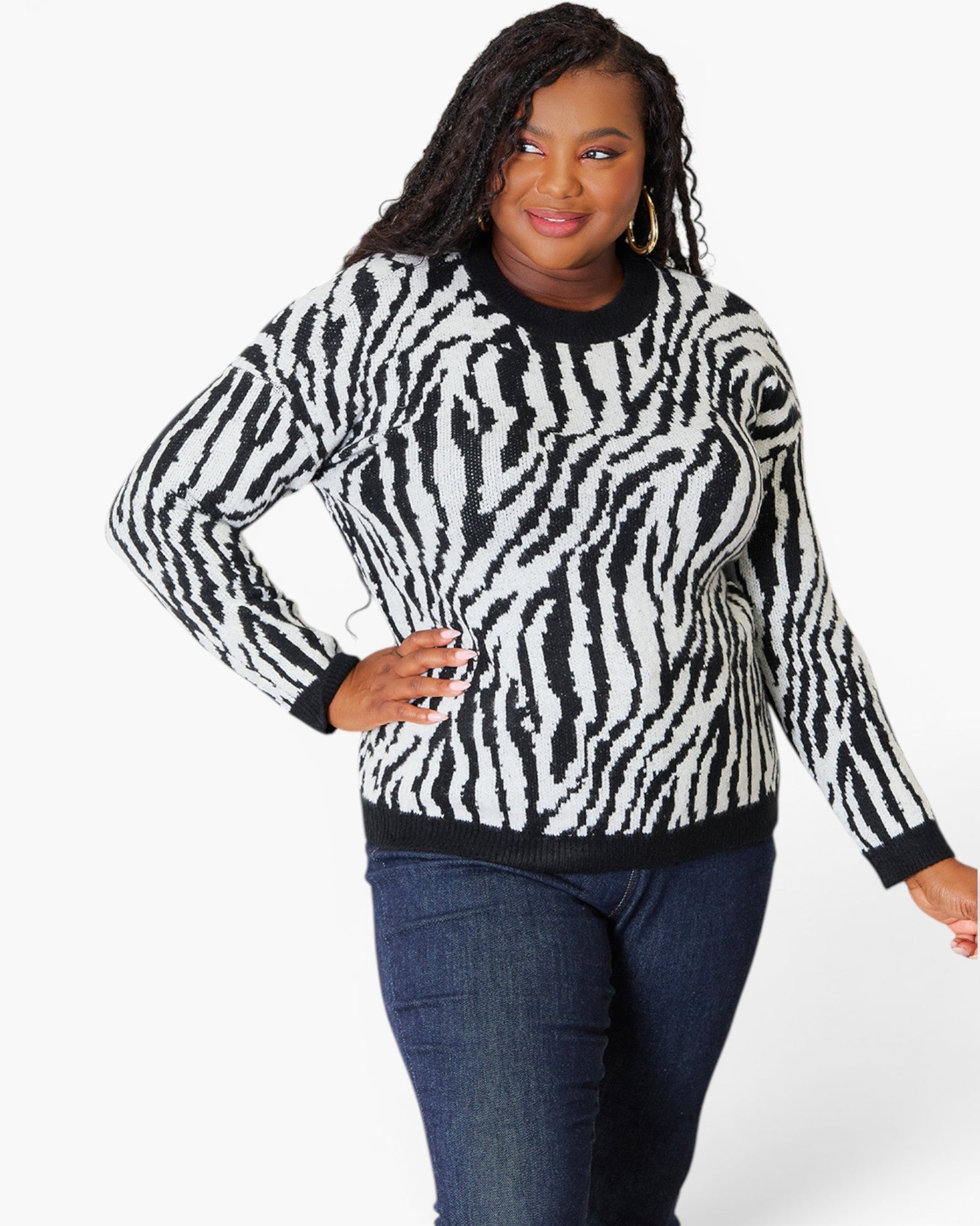 A woman modeling a black and white zebra-print long-sleeve sweater with black ribbed trim, paired with dark blue jeans, against a plain white studio background.