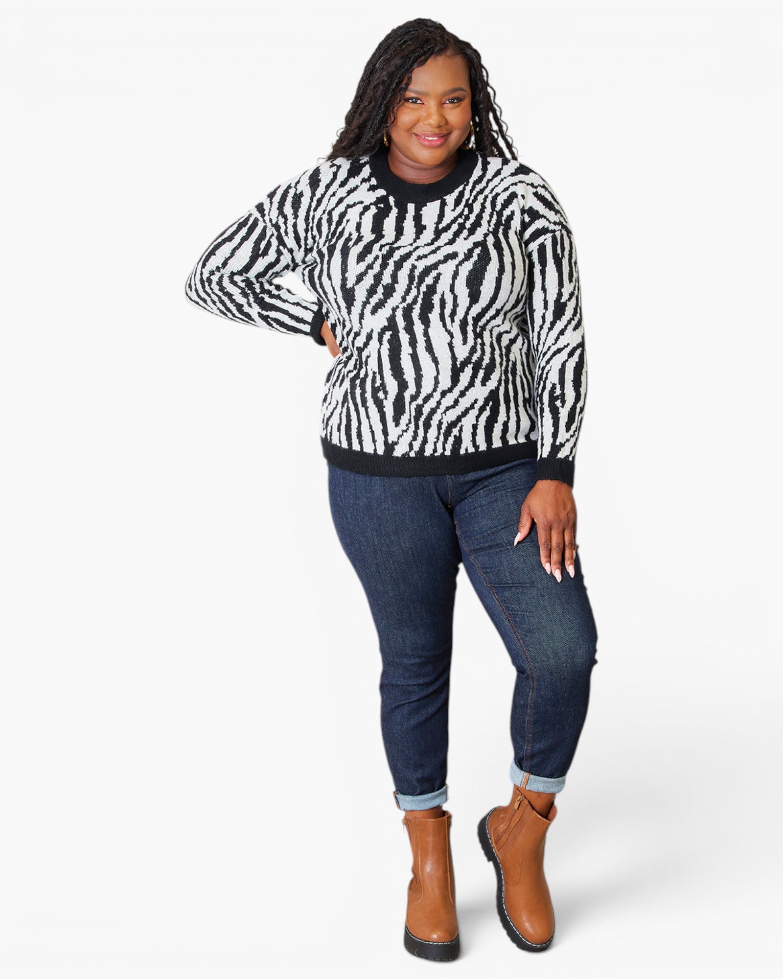 A woman modeling a black and white zebra-print sweater with dark blue jeans and brown ankle boots, standing against a light gray studio background.