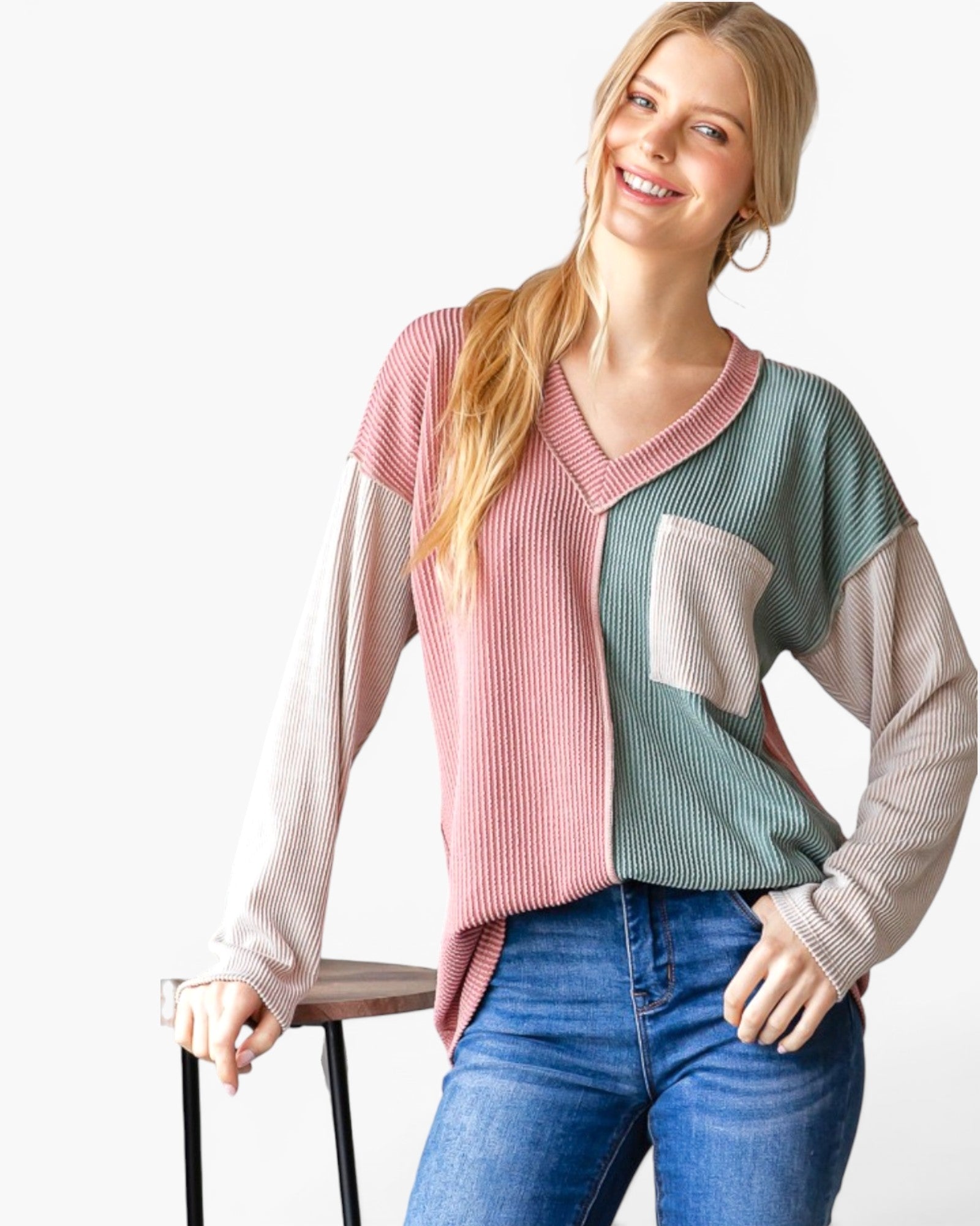 A woman wearing a pastel color-block ribbed knit sweater with pink, sage green, and beige sections, paired with blue jeans, standing against a white studio background.