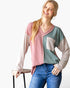 A woman wearing a pastel color-block ribbed knit sweater with pink, sage green, and beige sections, paired with blue jeans, standing against a white studio background.