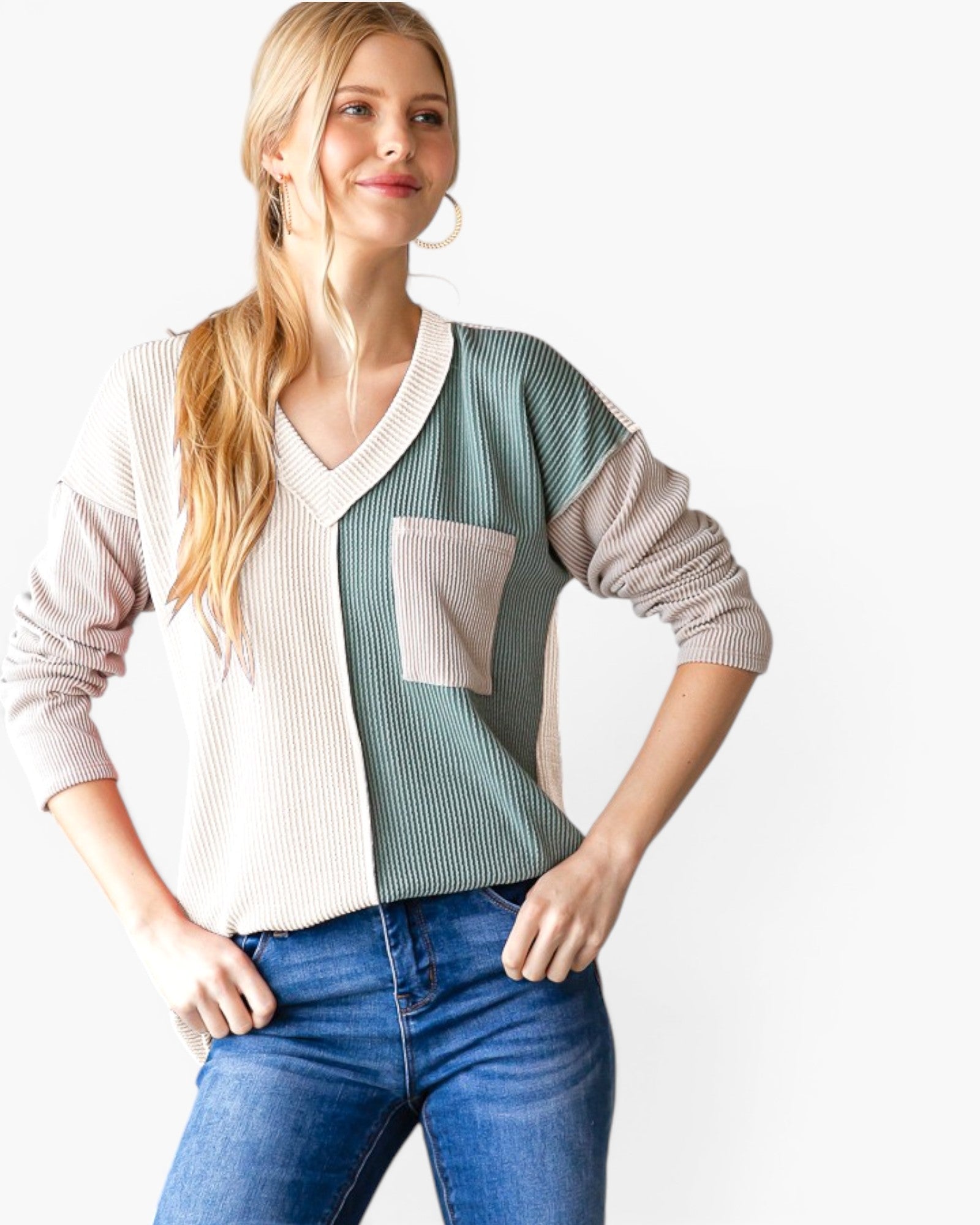 A woman modeling a color-block ribbed knit sweater with beige, sage green, and gray panels, paired with blue jeans against a plain white background.