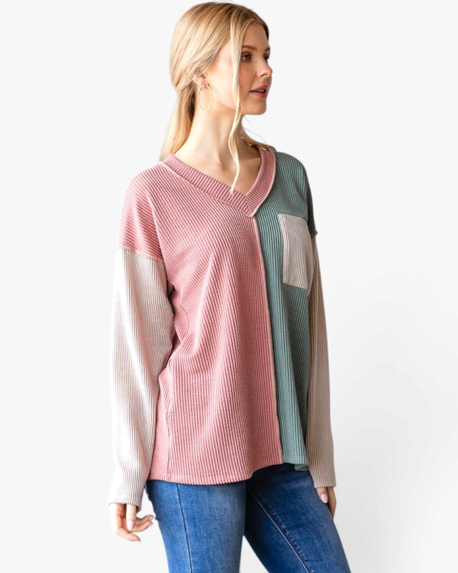 A woman wearing a color-blocked ribbed knit sweater with pink, sage green, and beige panels, paired with blue jeans, against a plain white background.