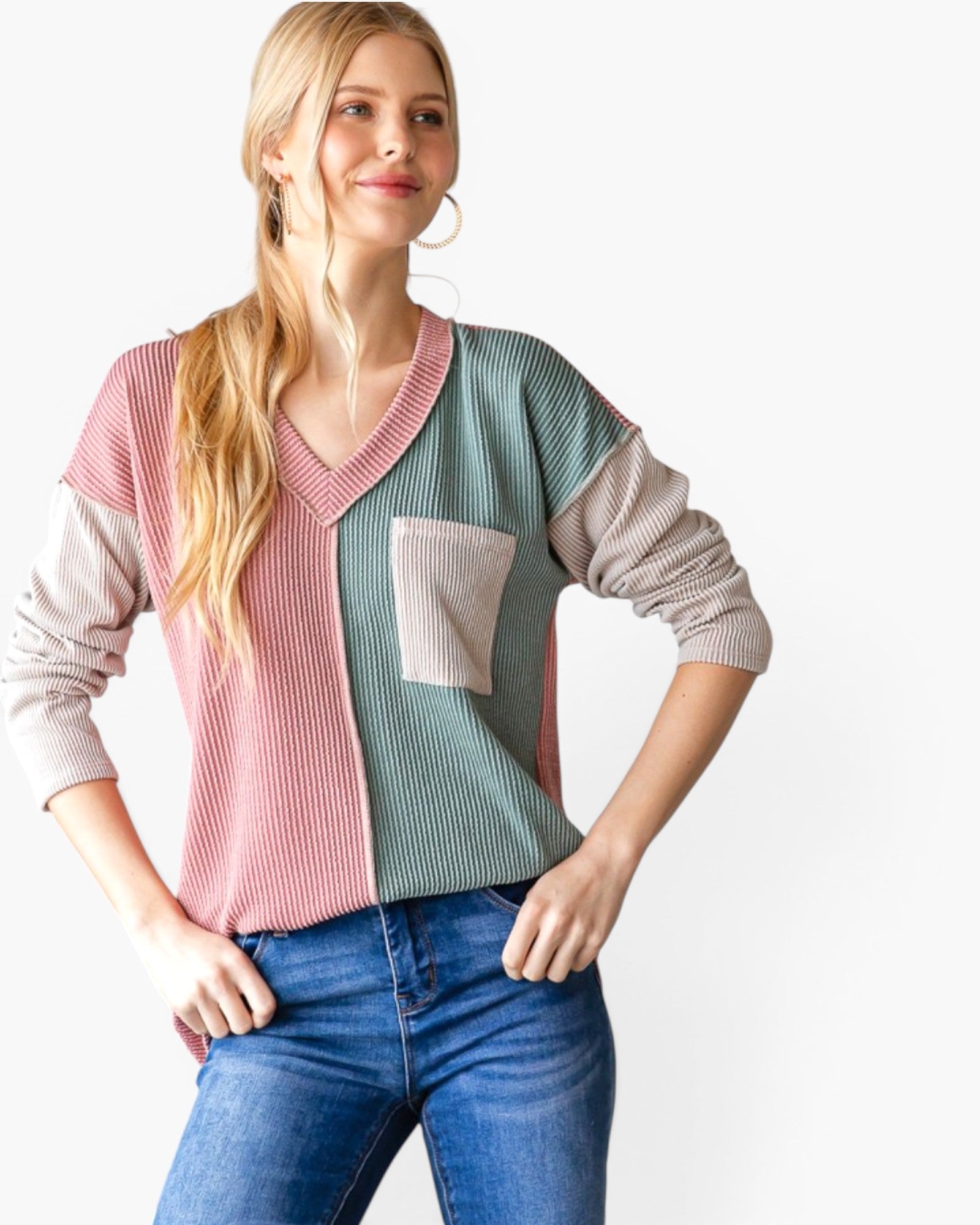 A woman modeling a color-blocked ribbed knit sweater with pink, sage green, and beige sections, paired with blue jeans against a plain white background.