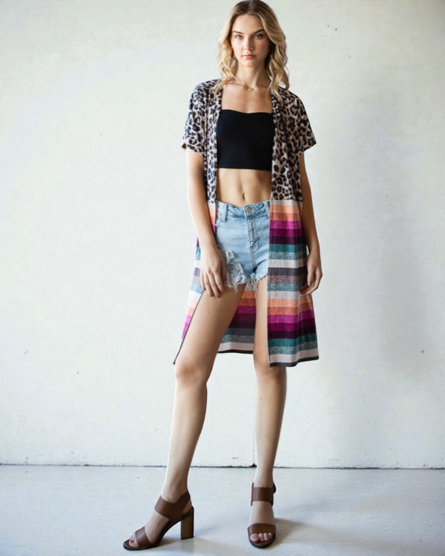 Woman wearing a leopard print shirt, black crop top, denim shorts, and a colorful skirt against a white background.