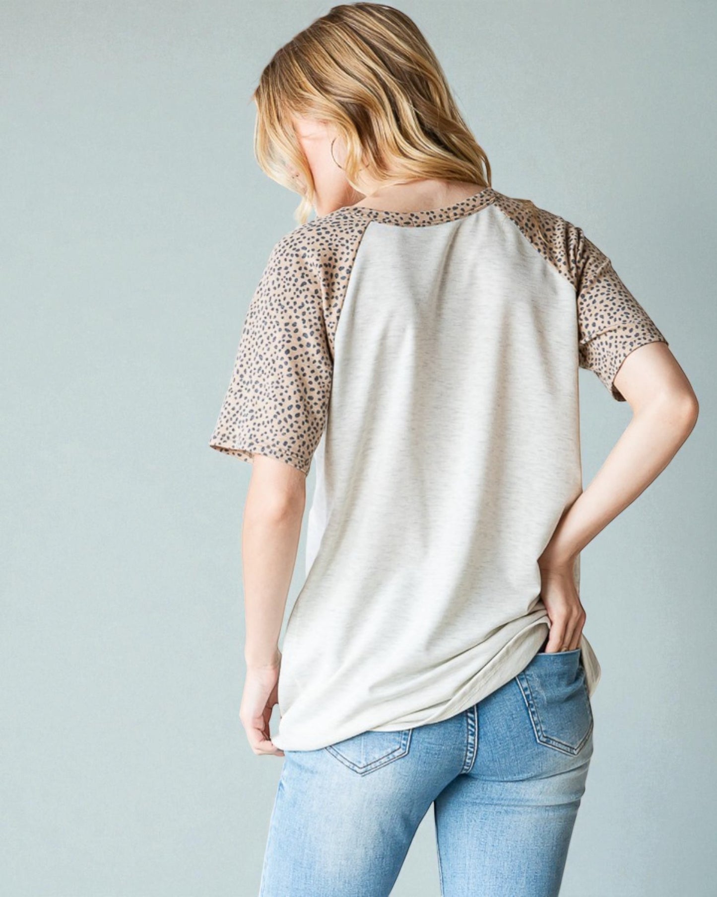 Woman wearing a beige t-shirt with leopard print sleeves and light blue jeans on a plain background